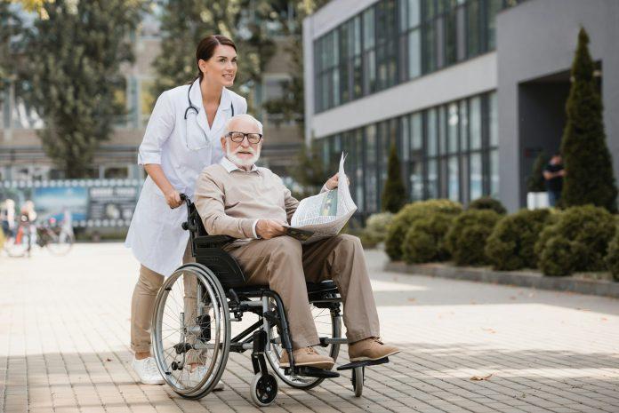 geriatric nurse walking with elderly disabled man in wheelchair with newspaper outdoors